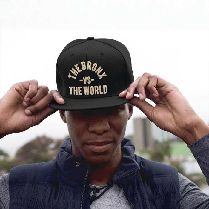 Black snapback hat with 'THE BRONX -VS- THE WORLD' in gold, worn by man in urban setting